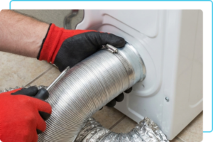 A professional wearing protective gloves carefully securing a metal transition hose to the back of a residential washing machine and dryer unit.